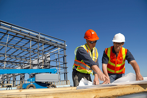 two men in orange safety vests and hard hats reviewing blue print as part of construction monitoring services